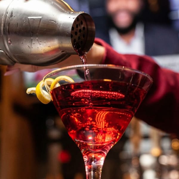 A bartender pours a red cocktail from a shaker into a martini glass with a lemon twist.
