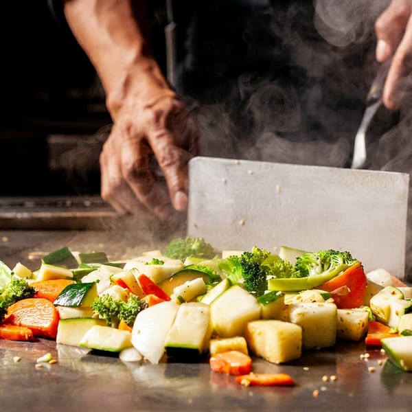 Chef cooking steaming vegetables like broccoli, carrots, and zucchini on a hibachi grill.