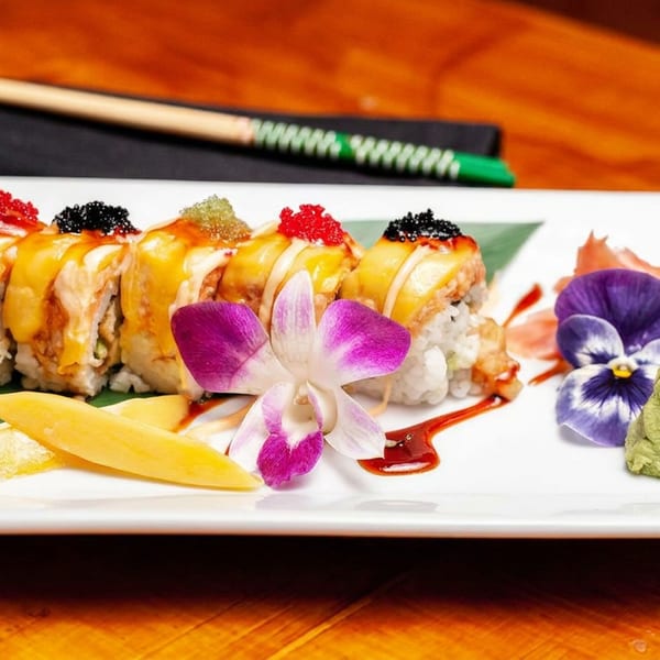 Sushi rolls topped with roe, mango slices, and edible flowers on a white plate with chopsticks.