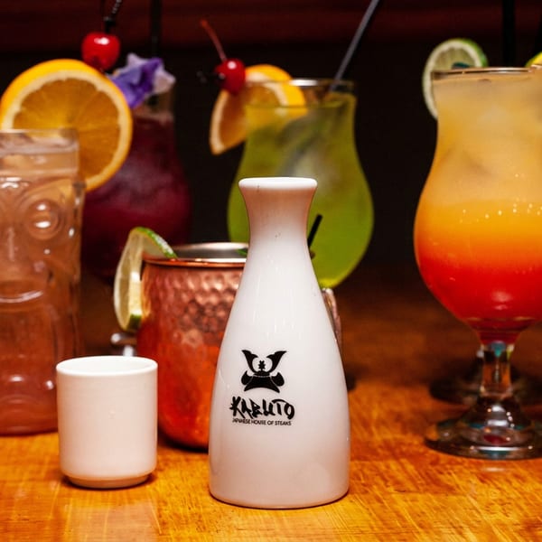A variety of colorful cocktails and a Kabuto sake bottle on a wooden bar.