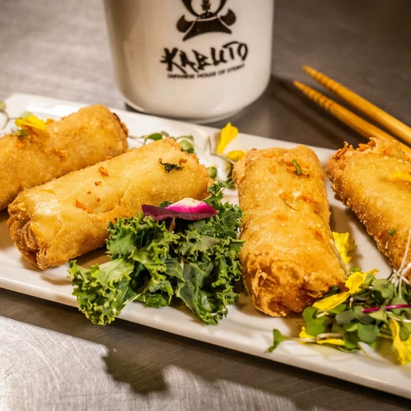 Four crispy golden spring rolls garnished with greens and edible flowers, with chopsticks and a branded cup.