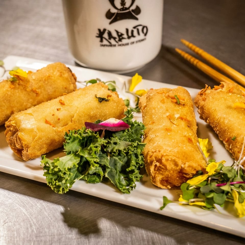 Four crispy golden spring rolls garnished with greens and edible flowers, with chopsticks and a branded cup.
